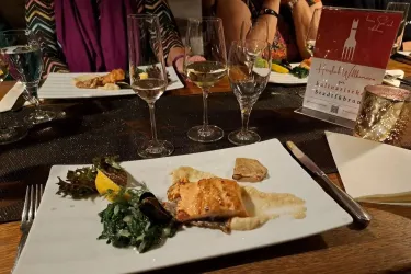 Ein schön angerichteter Teller mit gegrilltem Fisch und Gemüse. Im Hintergrund stehen Gläser mit Wein und man sieht andere Gäste am Tisch.