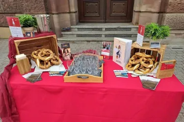 Ein Tisch mit frischen Brezeln und verschiedenen Holzbehältern steht vor einer historischen Fassade. Die dekorative Präsentation umfasst auch Informationsmaterialien über eine Veranstaltung.