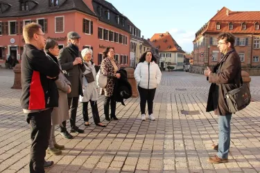 Eine Gruppe von Touristen hört einem Stadtführer auf einem Platz zu. Im Hintergrund sind historische Gebäude und ein klarer Himmel zu sehen.
