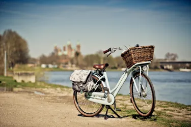 Ein schönes Fahrrad mit einem Korb steht am Ufer eines Flusses. Im Hintergrund sind Bäume und eine historische Architektur zu sehen.