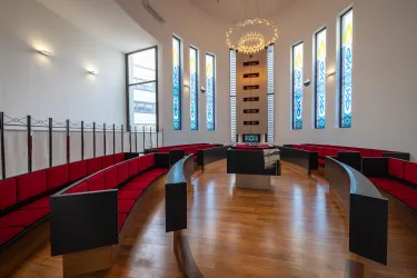 Ein moderner Innenraum mit roten Sitzbänken und großen Fenstern. Das Ambiente ist hell und freundlich, mit einem eleganten Kronleuchter in der Mitte.