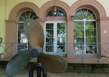 Ein Hubschrauberpropeller steht vor einem Gebäude mit großen Fenstern und einem Schild, das "Museum im Airlebnisraum" zeigt. Das Gebäude hat eine elegante Architektur und ist von Bäumen umgeben.