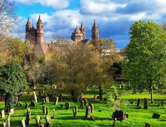 Ein historischer Friedhof mit vielen alten Grabsteinen und üppigem Grün. Im Hintergrund sind beeindruckende Türme eines Gebäudes und ein blauer Himmel zu sehen.
