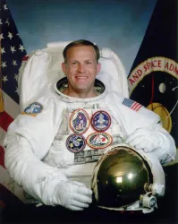 Ein Astronaut in einem Raumanzug lächelt in die Kamera. Im Hintergrund sind Flaggen und Embleme zu sehen.