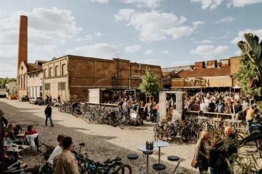 Ein lebhaftes Straßenfest mit vielen Menschen und Fahrrädern vor einer alten Fabrik. Der sonnige Himmel und die entspannte Atmosphäre laden zum Verweilen ein.