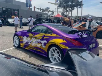 Ein auffälliges lila Auto mit einer bunten Folierung steht auf einem Parkplatz. Im Hintergrund sind weitere Fahrzeuge und Menschen auf einem Auto-Event zu sehen.