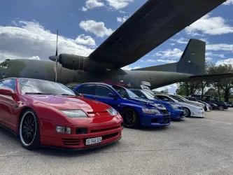 Eine Reihe von sportlichen Autos steht vor einem großen Flugzeug. Der Himmel ist klar mit einigen Wolken.