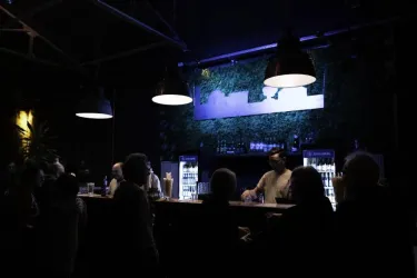 Eine stilvolle Bar mit sanfter Beleuchtung und einer grünen Wanddekoration. Menschen stehen am Tresen und unterhalten sich.