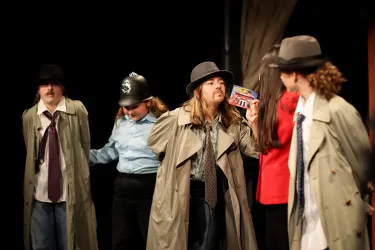 Eine Gruppe von Schauspielern in Kostümen steht auf der Bühne. Sie tragen Mäntel und Hüte und wirken wie Detektive oder Kriminelle in einer Theateraufführung.