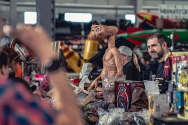 Ein Marktstand mit einem großen ET-Plüschtier und mehreren Sammelobjekten. Im Hintergrund sind Menschen und verschiedene Artikel zu sehen.