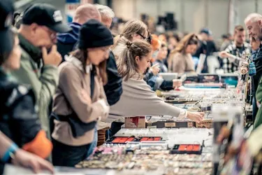 Eine Gruppe von Menschen an einem Marktstand, die verschiedene Produkte betrachten. Die Atmosphäre wirkt lebhaft und geschäftig.