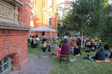 Eine Gruppe von Menschen sitzt im Garten vor einem roten Gebäude. Sie sind unter einem Zelt versammelt und scheinen einer Veranstaltung zuzuhören.