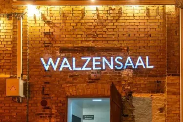 Ein Gebäude mit einer Backsteinfassade und einer beleuchteten Schrift, die "WALZENSAAL" zeigt. Der Eingang ist sichtbar und es ist eine gemütliche Atmosphäre zu erkennen.