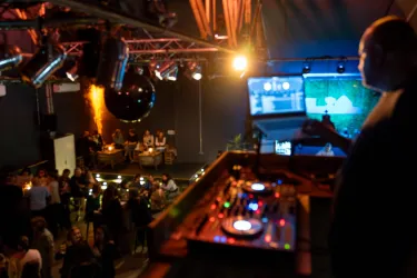 Eine belebte Lounge mit Menschen, die sich entspannen und tanzen. Im Vordergrund steht ein DJ an einem Mischpult mit bunten Lichtern.