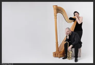 Ein Musiker sitzt mit einem Saxophon auf einem Hocker, während eine Harfenistin hinter ihm steht. Beide tragen elegante schwarze Kleidung und posieren in einem minimalistischen Hintergrund.