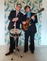 Zwei Musiker in Anzügen stehen vor einem bunten Hintergrund. Einer spielt auf einer Trommel und der andere hält eine Gitarre.