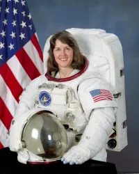Eine Astronautin in einem Raumanzug steht vor einer US-Flagge. Sie lächelt und hält einen Helm in der Hand.