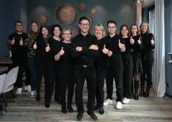 Eine Gruppe von Mitarbeitern posiert fröhlich in einem Raum und zeigt Daumen hoch. Alle tragen schwarze Kleidung und lächeln in die Kamera.