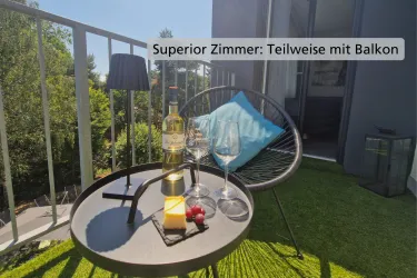 Ein gemütlicher Balkon mit einem schwarzen Tisch, zwei Weingläsern und einer Flasche Wein. Auf dem Tisch liegt ein Stück Käse und Trauben, umgeben von grüner Rasenfläche.