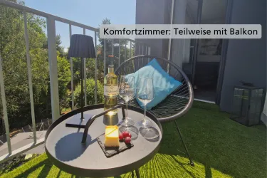 Ein gemütlicher Balkon mit einem runden Tisch, auf dem eine Flasche Wein, Gläser und ein Käseteller stehen. Der Schaukelstuhl und der grüne Rasenbelag schaffen eine einladende Atmosphäre.