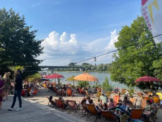 Ein lebhafter Ort am Wasser mit Liegen und bunten Sonnenschirmen. Menschen genießen die Sonne und die entspannte Atmosphäre.