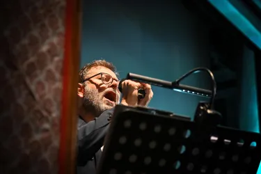 Ein Mann singt leidenschaftlich ins Mikrofon. Er steht vor einem Notenständer mit beleuchtetem Text.