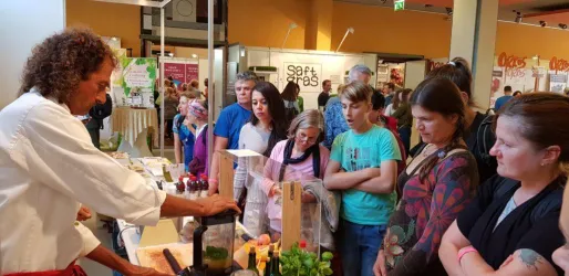 Ein Koch demonstriert die Zubereitung von Speisen vor einem interessierten Publikum. Viele Zuschauer schauen gespannt zu und nehmen Notizen.