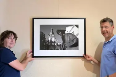 Zwei Personen halten ein gerahmtes Schwarz-Weiß-Foto entlang einer Wand. Das Bild zeigt eine beeindruckende Architektur und eine Figur im Vordergrund.