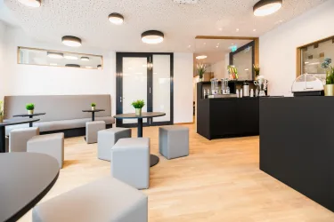 Ein modernes Café mit hellen Wänden und gemütlicher Einrichtung. Kleine Tische und quadratische Sitzblöcke laden zum Verweilen ein.