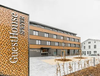 Ein modernes Hotelgebäude mit dem Namen "GuestHouse Speyer". Die Fassade ist in neutralen Farben gehalten und es gibt angelegte Grünflächen in der Umgebung.
