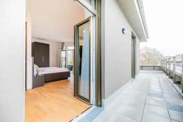 Ein moderner Balkon mit hellen Fliesen und Zugang zu einem Schlafzimmer. Das Zimmer ist gut beleuchtet und verfügt über große Fenster.