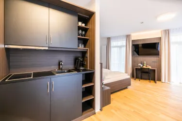 Eine moderne, helle Apartmentküche mit Holzboden und stilvollen Einbauschränken. Im Hintergrund ist ein gemütliches Schlafzimmer mit einem Fernseher zu sehen.