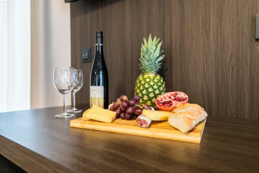 Eine rustikale Brett mit verschiedenen Käsesorten, Trauben, einer Ananas und einer Granatapfelhälfte. Im Hintergrund stehen zwei Weingläser und eine Flasche Wein.