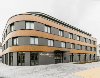 Ein modernes Gästehaus mit einer auffälligen Fassade aus Holz und Beton. Das Gebäude hat große Fenster und einen einladenden Eingangsbereich.