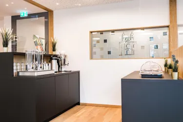 Ein modernes Foyer mit einem schwarzen Empfangstresen und einer attraktiven Kaffeetheke. Im Hintergrund ist ein großes Fenster, das eine stilvolle Transparenz bietet.