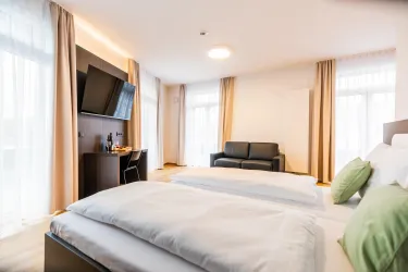 Ein modernes Hotelzimmer mit zwei Betten, einem Fernseher und einem Sitzbereich. Große Fenster sorgen für viel Tageslicht.