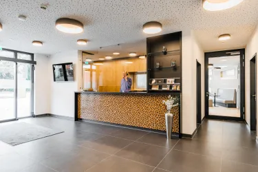 Eine moderne Hotelrezeption mit einer freundlichen Atmosphäre. Der Empfangstresen ist aus Holz gestaltet und ein Mitarbeiter steht bereit, um Gäste willkommen zu heißen.