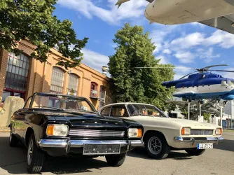 Zwei klassische Autos stehen vor einem historischen Gebäude. Im Hintergrund sind Bäume und ein Hubschrauber zu sehen.