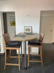 Ein kleiner Essbereich mit einem runden Tisch und zwei hohen Stühlen. Die Wände sind hell, und ein Kunstwerk hängt im Hintergrund.