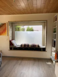 Ein gemütlicher Fensterplatz mit Kissen und einer Decke. Der Blick geht auf einen hellen Außenbereich mit Pflanzen.