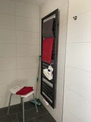 Ein modernes Badezimmer mit einem Handtuchheizkörper. Auf einem Hocker liegen rote und graue Handtücher sowie Reinigungsmittel.