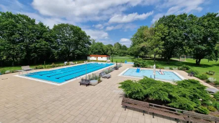 Ein Freibad mit einem großen Sportbecken und einem Kinderbecken. Umgeben von Bäumen und einer grünen Rasenfläche.