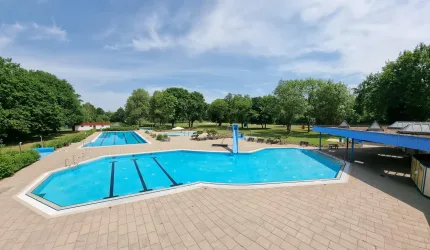 Ein Schwimmbad mit mehreren Becken und einem klaren blauen Himmel. Umgeben von grünen Bäumen und einer einladenden Umgebung.