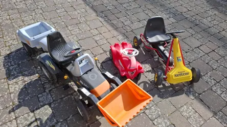 Drei Spielzeuge in Form von Fahrmaschinen stehen auf einem gepflasterten Boden. Ein Traktor, ein rotes Auto und ein gelber Kettcar sind gut sichtbar.