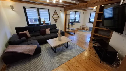 Ein modernes Wohnzimmer mit einem bequemen Sofa und einem Holzfußboden. Die großen Fenster lassen viel Licht herein und schaffen eine einladende Atmosphäre.