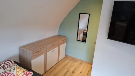 Ein gemütliches Zimmer mit einem Holzschrank und einem Spiegel. An der Wand hängt ein Fernseher, und der Boden ist aus hellem Holz.
