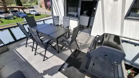 Ein moderner Balkon mit einem schwarzen Tisch und Stühlen. Ein Grill steht ebenfalls bereit für gemütliche Grillabende.