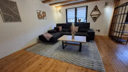 Ein modernes Wohnzimmer mit einer schwarzen L-förmigen Couch und einem Holzfußboden. Der Raum ist geschmackvoll dekoriert mit Kunstwerken an den Wänden und einem gemütlichen Teppich.