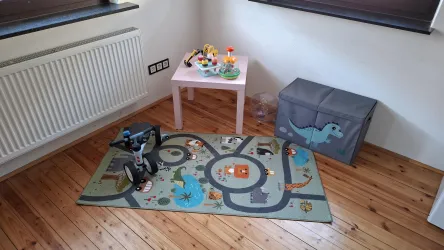 Ein kinderfreundliches Spielzimmer mit einem bunten Teppich und Spielzeug auf einem kleinen Tisch. In der Ecke steht eine graue Aufbewahrungsbox mit einem Dinosauriermuster.