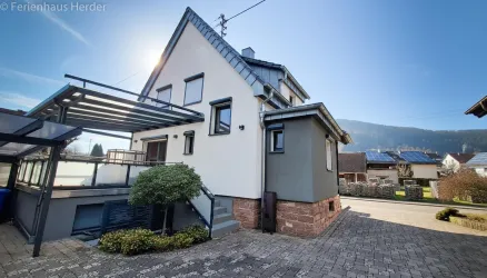 Ein modernes Haus mit einer Terrasse und einer klaren Fassade. Im Hintergrund sind sanfte Hügel und ein blauer Himmel zu sehen.